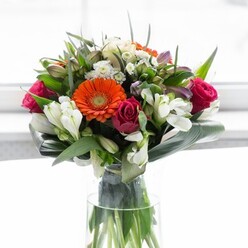 Anerley's charm is reflected in every nuance of this magnificent floral arrangement. At its heart are creamy peach and spirited orange gerbera daisies, their petals radiating outward like bursts of sunlight through the leafy canopy of Betts Park. Rich magenta roses provide a sumptuous counterpoint, their velvet petals swirling with romance and depth, while elegant ivory alstroemeria and fine white blossoms contribute layers of delicate lightness. Leafy aspidistra is wrapped protectively around the base, forming a lush green necklace that grounds the exuberant colours above, reminiscent of the calm found along the edges of Crystal Palace Park. Smaller green leaves and airy filler sprigs weave between the larger blooms, giving the arrangement natural dimension and a sense of quiet abundance. Presented in a tall, clear glass vase, the intertwined stems stand as a testament to the bouquet's vitality and skill of its local creator. Bathed in soft white light, each bloom's individual beauty shines, making this ensemble perfect for both joyful celebrations and heartfelt messages across Anerley and its neighbouring communities. The bouquet's artistry embodies optimism, gratitude, and timeless style.